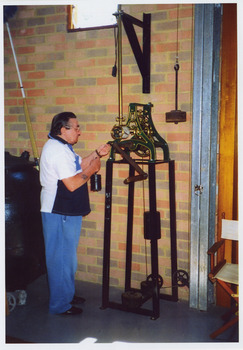 Shows a man oiling the clock winding mechanism in The Marysville Museum in Marysville in Victoria. The mechanism is painted green and gold and is mounted on a metal frame with a crank handle.