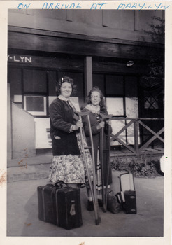A black and white photograph taken at Mary-Lyn Guest House in Marysville. Mary-Lyn was a part of the Mary Chain of guest houses built by Eric Dowdle in the Depression years. The photograph is of two ladies standing at the front of the main building with their luggage and each is holding a crutch.