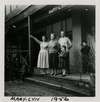 A black and white photograph taken at Mary-Lyn Guest House in Marysville. Mary-Lyn was a part of the Mary Chain of guest houses built by Eric Dowdle in the Depression years. The photograph was taken in 1956 and shows two ladies, a man and two children standing on the front verandah at Mary-Lyn.