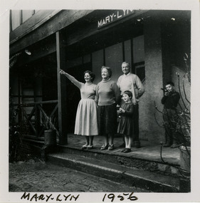 A black and white photograph taken at Mary-Lyn Guest House in Marysville. Mary-Lyn was a part of the Mary Chain of guest houses built by Eric Dowdle in the Depression years. The photograph was taken in 1956 and shows two ladies, a man and two children standing on the front verandah at Mary-Lyn.