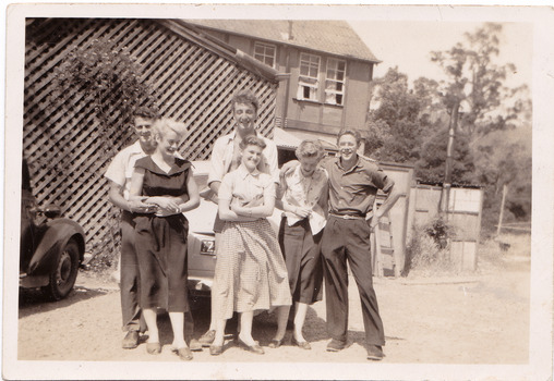 A black and white photograph taken at Mary-Lyn Guest House in Marysville. Mary-Lyn was a part of the Mary Chain of guest houses built by Eric Dowdle in the Depression years. The photograph was taken between 1954 and 1955 and is of an unknown group of people standing outside Mary-Lyn. The group of people may have worked at Mary-Lyn from 1954 to 1955.