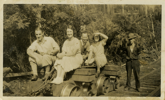 A black and white photograph of a group of guests who were staying at Mary-Lyn Guest House in Marysville. Mary-Lyn was a part of the Mary Chain of guest houses built by Eric Dowdle in the Depression years. The photograph, which was taken in 1933, shows a group of men and women sitting on top of a timber tramway logging jinker.
