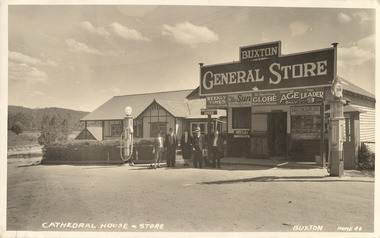 Shows the view from across the road towards a small house beside the Buxton General Store. Shows a weatherboard building with parapet at roof with lettering: Buxton General Store, signs for newspapers in the windows, two petrol bowsers on the path at each end of store. Shows a small group of people standing on the path between the house and store. The title of the postcard is hand written in white ink along the lower edge.