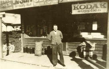 Shows a man standing outside the Buxton General Store. Shows advertising signs for Castrol fuel and Kodak photography supplies.