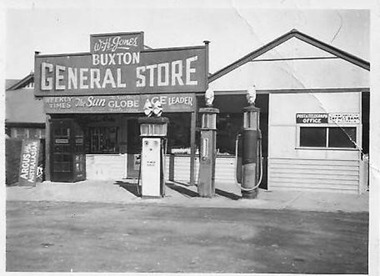 Shows the front facade of the Buxton General Store with the adjacent Post Office. Shows a large sign above the front of the store with the proprietor's name and the name of the store. There are also advertising signs for various newspapers. In front of the store are three petol bowsers.