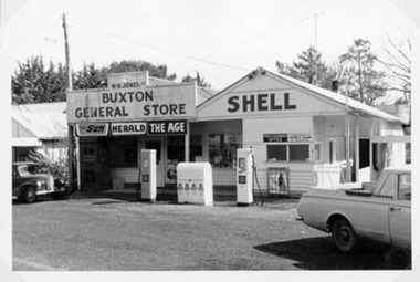 Shows the Buxton General Store in Victoria. Above the door is a large sign with the proprietor's name and the name of the store. There are three advertising signs for different newspapers. As well there is an advertising sign for Shell fuel. Adjacent to the store building is the Buxton Post Office building. Out the front of the post office building are two petrol bowsers. There are two cars parked outside the store. 