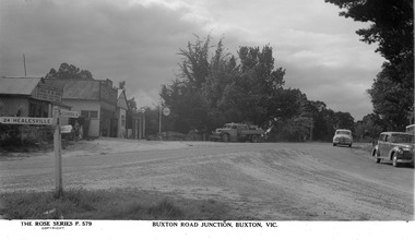 Shows the junction of two roads at Buxton in Victoria. Shows the Buxton General Store in the background with the Buxton Post Office adjacent to it at the right. To the left of the store is the Grandview Cafe which offers grills at all hours. There is a truck parked outside the store and there are two cars turning onto the Marysville-Buxton Road. In the foreground is a signpost with signs showing the distance and direction of Alexandra, Marysville and Healesville. 