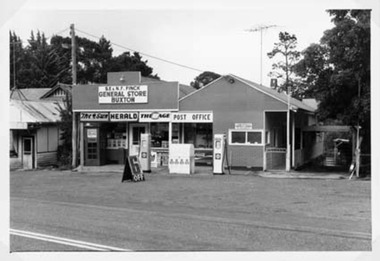 Shows the Buxton General Store in Victoria. Above the doorway is a sign with the then owners name and the name of the store. There are also advertising signs for three newspapers. The Post Office building is adjacent with two petrol bowsers out the front. 