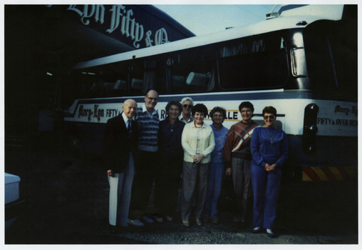 A copy of a colour photograph taken at Mary-Lyn '50 & Over Holiday Resort in Marysville. Mary-Lyn was a part of the Mary Chain of guest houses built by Eric Dowdle during the Depression years. The photograph shows a group of people standing in front of the bus used by the resort to transport its guests on various tours in the area.