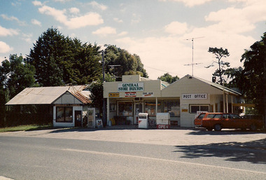 Shows the Buxton General Store in Victoria. Above the doorway is a sign with the name of the store. There are also advertising signs for three newspapers. The Post Office building is adjacent with one petrol bowser out the front. There is also a telephone paybox outside the store.