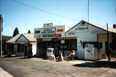 Shows the Buxton General Store in Victoria. Above the doorway is a sign with the name of the store. There are also various advertising signs for different goods and services including newspapers. There is a red Post Office box out the front with one petrol bowser next to it. There is also a telephone paybox outside the store.