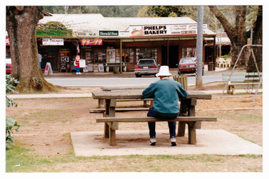 Shows the front facade of Phelps Bakery in Marysville in Victoria. Shows the bakery on the corner with the newsagent next door. In the foreground of the photograph there is a wooden table with bench seats with a man seated at the table.