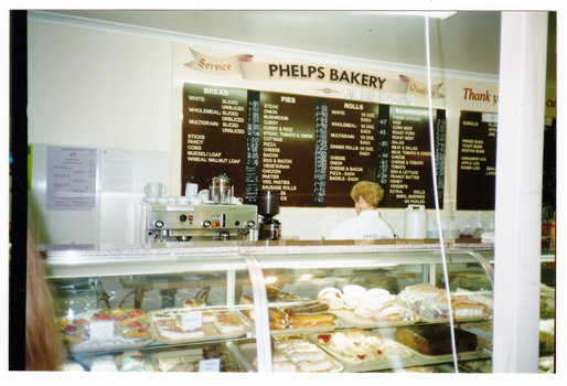 Shows the front counter in Phelps Bakery in Marysville in Victoria. Shows a wall mounted menu of some of the food that is available and shows a glass fronted cabinet with some of the baked goods on offer to customers.