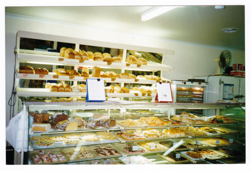 Shows the front counter in Phelps Bakery in Marysville in Victoria. Shows a glass fronted cabinet with some of the baked goods on offer to customers. Above the counter are shelves with various types of loaves of bread. In the background there is a pie warmer.