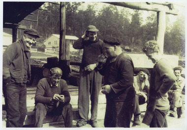 A copy of a black and white photograph taken at 'smoko' at Goulds Sawmill in Marysville. The original photograph was taken in 1950-1960 and shows a group of men standing around enjoying their smoko break.