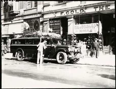 Shows a black and white photograph of the Marysville Tourist Bureau in Melbourne. Shows the building with a the bus waiting out the front to take passengers from Melbourne to Marysville. Shows a man, possibly the driver, polishing the bus. 