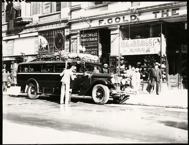 Shows a black and white photograph of the Marysville Tourist Bureau in Melbourne. Shows the building with a the bus waiting out the front to take passengers from Melbourne to Marysville. Shows a man, possibly the driver, polishing the bus. 