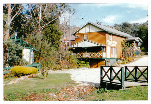 Shows a photograph of the main building and entrance gate at Arbor Green Galleries in Marysville. Shows a large brick building with a green and cream painted timber arbor. In the foreground is a small green painted timber bridge.
