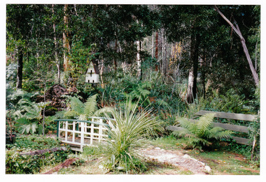 Shows a photograph of the rear garden at Arbor Green Galleries in Marysville. Shows a small timber bridge with a bird house standing next to it.