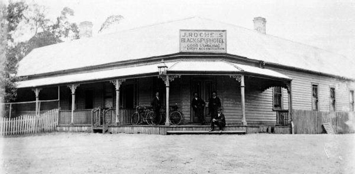 Shows a black and white photograph of J.Roches Black Spur Hotel in Narbethong. Shows a weatherboard building with a verandah running along the front facade. Shows a group of four men standing near the entrance door alongside a group of bicycles.