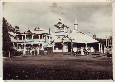Shows a black and white photograph of the Black Spur Hotel and Narbethong House in Narbethong. The Black Spur Hotel is a weatherboard building with a verandah running along the front and left side of the building. Narbethong House is a double storey weatherboard building with verandahs on both levels. There is a set of stairs leading up to the front entrance. There is a kangaroo and a kookaburra carved from wood on the gabled peaks of the roof of Narbethong House. There is a group of men standing on the verandah of Narbethong House as well as two men standing on the verandah of the Black Spur Hotel. In front of the hotel is an early model car with a man and women standing either side of the car and another man standing at the rear of the car.