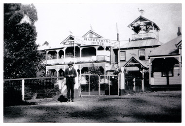 Shows a black and white photograph of Narbethong House situated next to the Black Spur Hotel in Narbethong. Narbethong House is a double storey weatherboard building with verandahs on both levels. There is a wood carved kangaroo, kookaburra, dingo and emu on the gabled peaks of the roof. In front of the gate leading into Narbethong House there is a man standing posing for the photograph.
