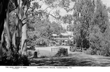 Shows a black and white photograph of Narbethong House situated next to the Black Spur Hotel in Narbethong. Narbethong House is a double storey weatherboard building with verandahs on both levels. There is a wood carved kangaroo and kookaburra on the gabled peaks of the roof. In the grounds in front of Narbethong House are a number of early model cars.