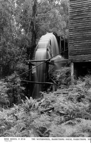 Shows a waterwheel situated next to a wooden building. There is water going over the waterwheel which is surrounded by bushland.