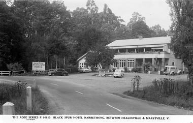Shows a black and white postcard of the Black Spur Hotel in Narbethong. Shows a large double storey building with a verandah running along the front of both floors. Shows several vehicles standing in the front car part of the hotel alongside a sign advertising counter lunches 12 to 2pm daily and sign advertising the availability of Melbourne Bitter and Penfolds wine at the hotel.