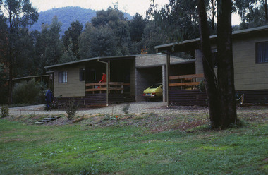 Shows two of the cottages located at Blackwood Cottages in Marysville. Shows two weatherboard buildings both with small verandahs at the front. There is a lady standing on the verandah of one of the cottages with a car under the carport. The cottages are both surrounded by large trees.
