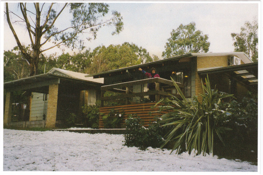 Shows two of the cottages located at Blackwood Cottages in Marysville. Shows two buildings, one with a small verandah at the front. There is a lady standing on the verandah of one of the cottages. There is snow on the ground in front of the cottages.