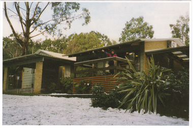 Shows two of the cottages located at Blackwood Cottages in Marysville. Shows two buildings, one with a small verandah at the front. There is a lady standing on the verandah of one of the cottages. There is snow on the ground in front of the cottages.