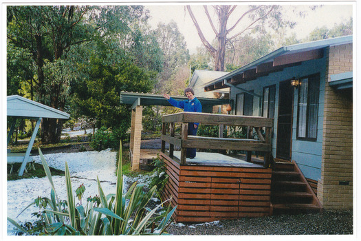 Shows two of the cottages located at Blackwood Cottages in Marysville. Shows two buildings, one with a small verandah at the front. There is a lady standing on the verandah of one of the cottages. There is snow on the ground in front of the cottages.