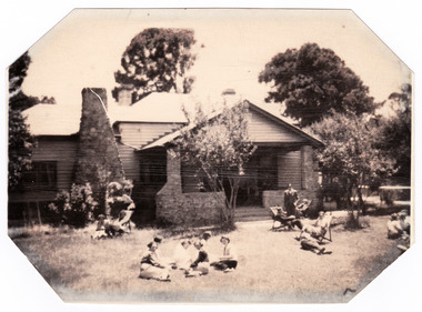 Shows a weatherboard farmhouse with a large stonework chimney and stonework porch at the front of the house. There are several people sitting on chairs and on the front lawn of the house. 