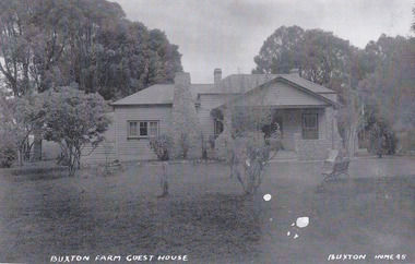 Shows a weatherboard farmhouse with a large stonework chimney and stonework porch at the front of the house. There are a few benches and seats arranged on the front lawn.