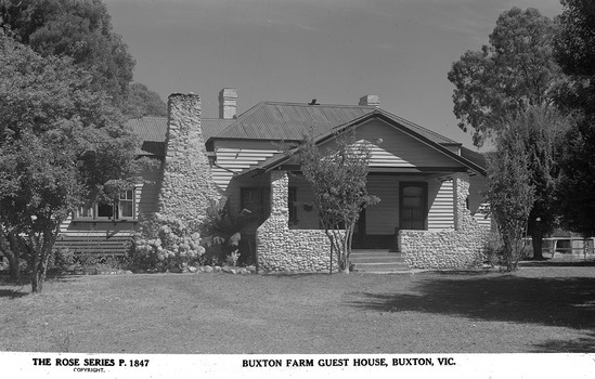 Shows a weatherboard farmhouse with a large stonework chimney and stonework porch at the front of the house.