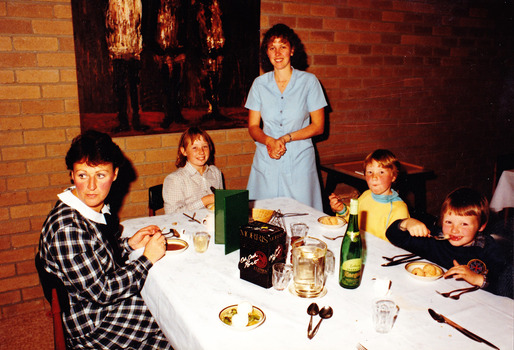 A digital copy of a colour photograph of a woman and three young children eating dinner at Mountain Lodge with a young girl standing looking on. The photograph was taken in 1985. 