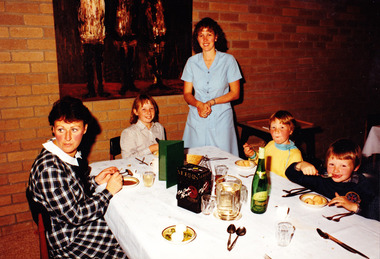 A digital copy of a colour photograph of a woman and three young children eating dinner at Mountain Lodge with a young girl standing looking on. The photograph was taken in 1985. 