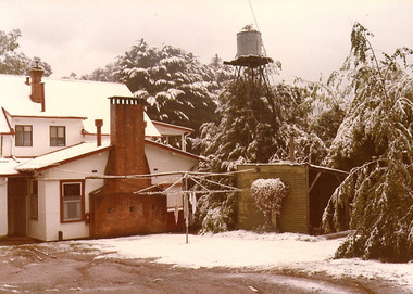 A copy of a sepia photograph taken at Kooringa Tourist House in Marysville. The photograph is of the main building after a snowfall. Photograph also shows a rotary washing line with a raised water tank in the background.
