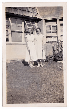 A black and white photograph taken at Mary-Lyn Guest House in Marysville. Mary-Lyn was part of the Mary Chain of guest houses built by Eric Dowdle in the Depression years. The photograph shows two ladies wearing white dresses and holding tennis rackets standing in front of the guest house.