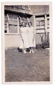 A black and white photograph taken at Mary-Lyn Guest House in Marysville. Mary-Lyn was part of the Mary Chain of guest houses built by Eric Dowdle in the Depression years. The photograph shows two ladies wearing white dresses and holding tennis rackets standing in front of the guest house.