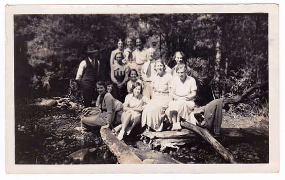 A black and white photograph taken of a group of guests who were staying at Mary-Lyn Guest House in Marysville. Mary-Lyn was part of the Mary Chain of guest houses built by Eric Dowdle in the Depression years. The photograph shows the group of thirteen people sitting on logs in the forest at an unknown location.
