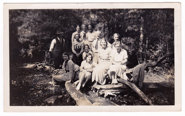 A black and white photograph taken of a group of guests who were staying at Mary-Lyn Guest House in Marysville. Mary-Lyn was part of the Mary Chain of guest houses built by Eric Dowdle in the Depression years. The photograph shows the group of thirteen people sitting on logs in the forest at an unknown location.