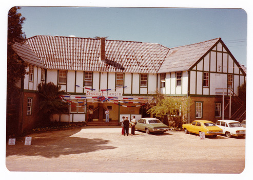 A colour photograph taken at Mary-Lyn Guest House in Marysville. Mary-Lyn was a part of the Mary Chain of guest houses built by Eric Dowdle in the Depression years. The photograph shows a view of the rear entrance of the main building with a banner above the door that says "Sterlings Tavern".