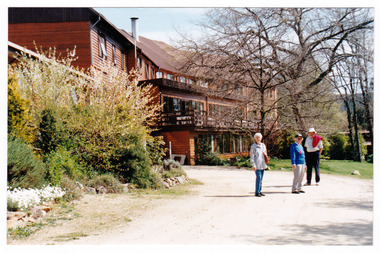 A colour photograph of Mary-Lyn Guest House in Marysville. Mary-Lyn was a part of the Mary Chain of guest houses built by Eric Dowdle during the Depression years. The photograph shows the rear view of the main building with a group of three adults standing in front of the building.