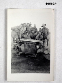 Photograph of a group of Australian soldiers in uniform, standing around a wrecked jeep.