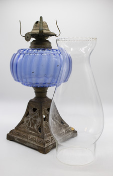 A lamp with embossed metal base and blue glass fuel bowl stands next to a clear glass lamp shade, placed in the foreground.