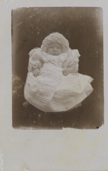 A postcard with a black-and-white image of a baby in a lace dress with a frilly hat.