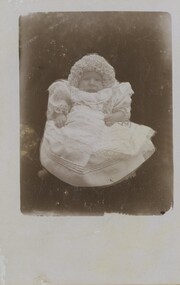 A postcard with a black-and-white image of a baby in a lace dress with a frilly hat.