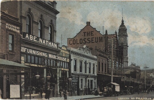 A row of early 1900s buildings along a street. There is a clocktower towards the end of the row and people walking along the footpath.